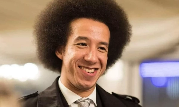 A smiling man with a large afro hairstyle, dressed in a black coat and tie, stands in a softly lit setting.