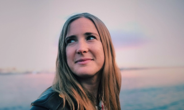 A young woman with long, straight hair gazes thoughtfully into the distance, with a serene water background and a soft colorful sky at sunset.