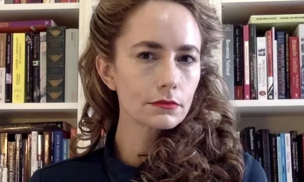 A woman with curly hair and red lipstick poses in front of a bookshelf filled with various books, exuding a confident expression.