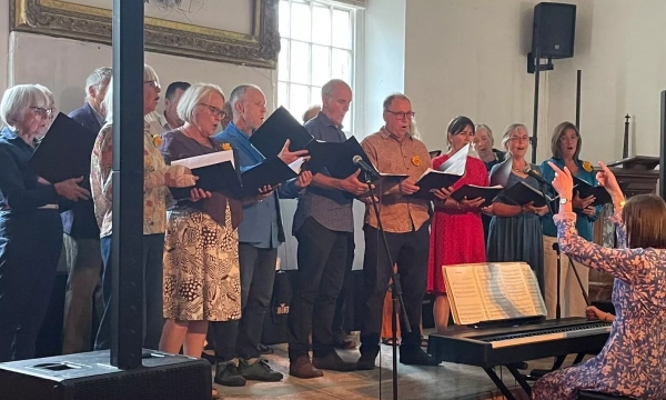 A diverse choir of men and women sings together, holding music sheets, with a conductor leading from the front near a piano.