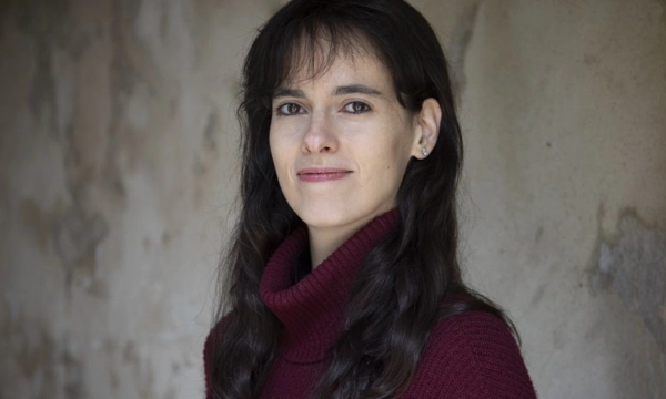 A woman with long, dark hair wearing a burgundy turtleneck sweater, standing against a textured, neutral background.