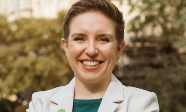 Smiling woman with short hair, dressed in a white blazer and green top, standing outdoors with greenery in the background.