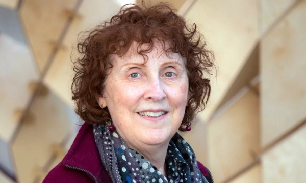 Image of a woman with curly, reddish-brown hair, wearing a maroon jacket and patterned scarf, smiling against a backdrop of geometric shapes.