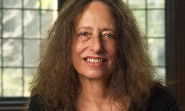 A woman with long, curly hair smiles warmly, seated in front of large windows with natural light, creating a serene atmosphere.