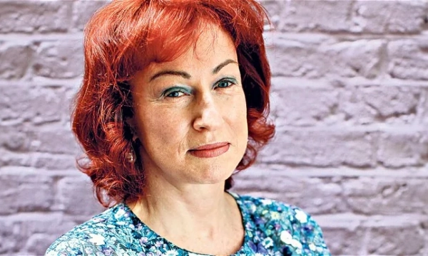 A woman with red hair and blue eye makeup poses against a textured gray wall, wearing a floral-patterned top.