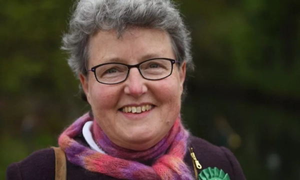 A smiling woman with gray hair and glasses, wearing a colorful scarf and a green rosette, stands outdoors with greenery in the background.
