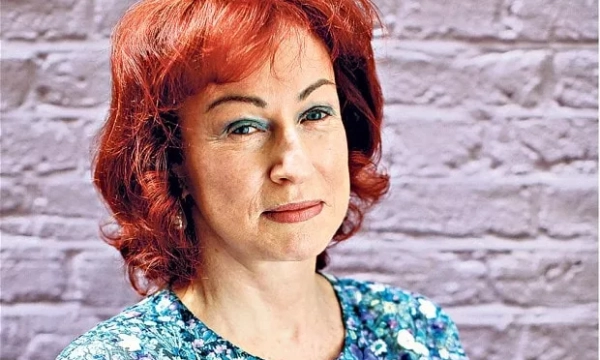 A woman with vibrant red hair poses against a textured, light-colored brick wall, wearing a floral-patterned top and a neutral expression.