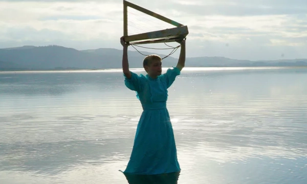 A person in a blue dress stands in shallow water, holding a triangular wooden frame overhead against a backdrop of serene lake and distant hills.