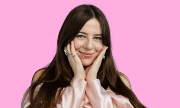 A person with long brown hair smiles and gently holds their face, wearing a pink satin top against a pink background.