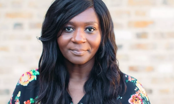 A woman with long, dark hair and a floral blouse is smiling softly against a brick wall background.