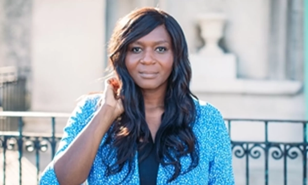 A woman with long, wavy hair wearing a blue patterned jacket stands outdoors, looking directly at the camera with a soft expression.