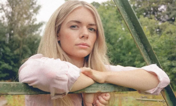 A young woman with long blonde hair leans against a green swing frame, displaying a serious expression amidst a natural, overgrown background.