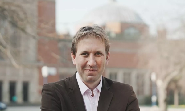 A smiling man in a brown blazer stands outdoors with a blurred, historic building in the background, suggesting an academic setting.