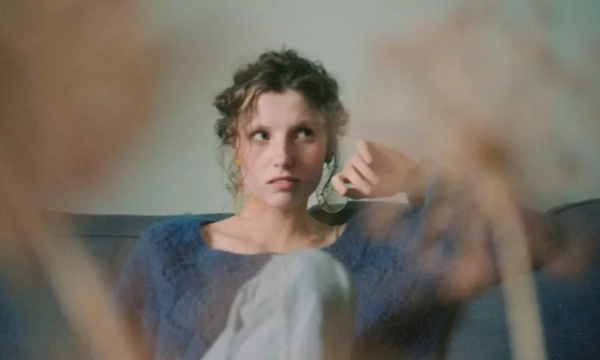 A young woman with curly hair sits on a blue couch, looking contemplatively while holding a cup, surrounded by out-of-focus plant stems.