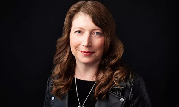 A woman with wavy brown hair and blue eyes wearing a black leather jacket, smiling against a dark background.