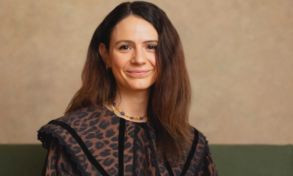 A woman with long dark hair, wearing a patterned blouse with ruffles, smiles while sitting against a textured background.