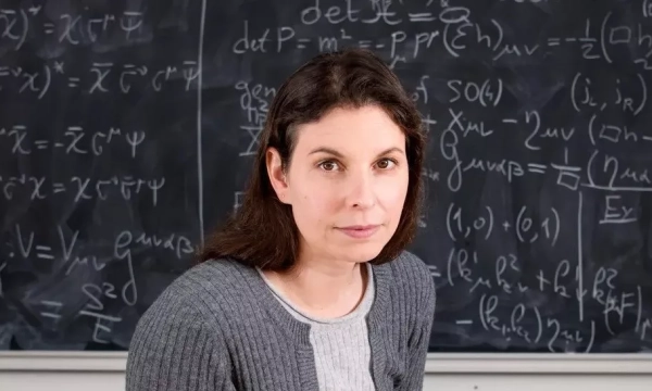 A woman with dark hair and a gray cardigan sits in front of a blackboard filled with mathematical equations and symbols.
