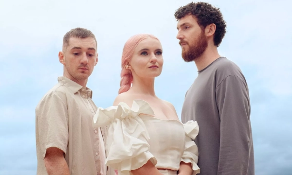 A trio of individuals stands outdoors against a cloudy sky, with one wearing a beige shirt, another in a white dress, and the last in a gray long-sleeve shirt.