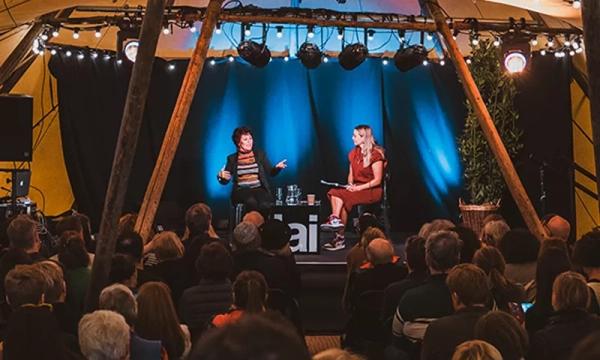 A speaker and moderator engage in conversation on stage, surrounded by an attentive audience under warm lights inside a tent-like venue.