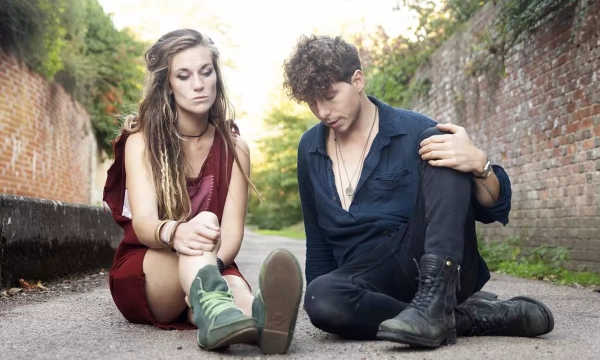 A young couple sits on a pathway, looking contemplative and slightly distressed, surrounded by greenery and brick walls.