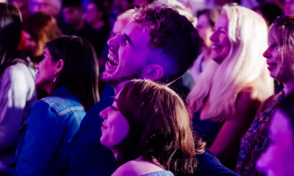A diverse group of people enjoying a lively event, with a focus on two individuals laughing and sharing a joyful moment in a colorful, vibrant setting.