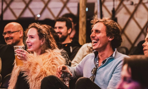 A group of people sitting together in a venue, laughing and enjoying drinks, with one person in a fluffy jacket.