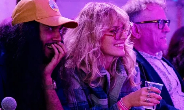 A group of three people smiling and engaged, with one wearing a yellow cap and another holding a cup, set against a purple-lit background.