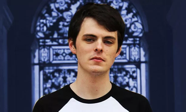 A young man with dark hair is posing in front of an ornate, patterned background, wearing a black and white shirt.