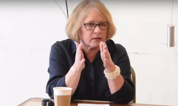 A woman with glasses gestures while seated at a table, discussing a topic with a coffee cup and notepad in front of her.