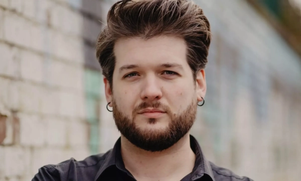 A young man with a beard and earrings stands in front of a textured brick wall, looking confidently at the camera.