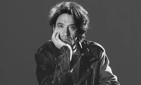 A man with tousled hair sits thoughtfully, resting his chin on his hand, wearing a black leather jacket against a dark background.