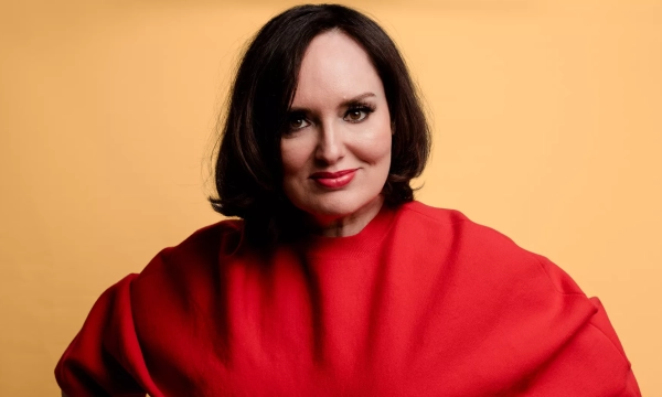 A smiling individual with shoulder-length dark hair, wearing a striking red garment, poses confidently against a yellow background.