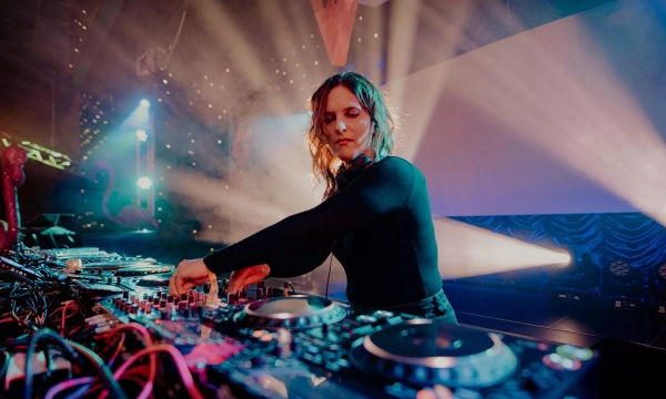 A female DJ performs at a vibrant event, surrounded by illuminated decks and colorful stage lights, creating an energetic atmosphere.