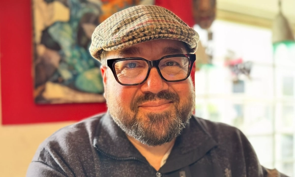 A man with a beard and glasses, wearing a flat cap, smiles warmly at the camera in a colorful cafe setting.