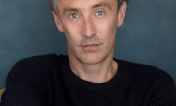 A portrait of a man with tousled hair and blue eyes, wearing a black sweater against a soft, muted background.