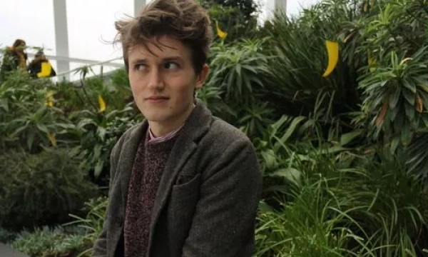 A young man with tousled hair sits among lush greenery, wearing a gray blazer and a cozy sweater, looking thoughtfully to the side.