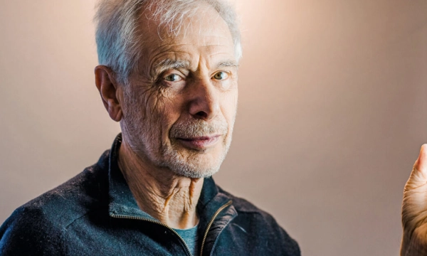 An older man with gray hair and a serious expression, holding a light source, set against a softly lit background.