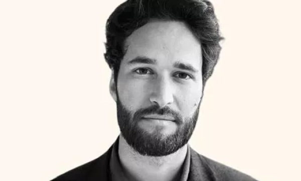 A close-up portrait of a man with a beard and tousled hair, wearing a dark blazer, against a plain light background.
