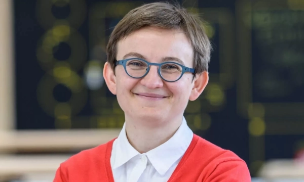 A person with short hair and glasses, wearing a red cardigan over a white shirt, smiles confidently in a bright, modern environment with abstract designs in the background.