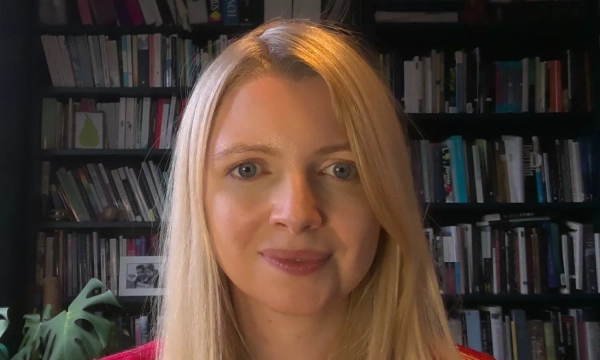 A woman with long blonde hair wearing a red top, standing in front of a bookshelf filled with various books.