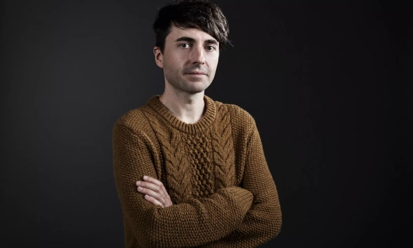 A man with dark hair, wearing a cozy brown cable-knit sweater, stands with arms crossed against a dark background.