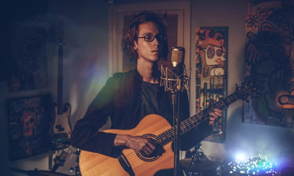 A young musician with glasses performs with an acoustic guitar in a dimly lit room adorned with colorful artwork and musical instruments.
