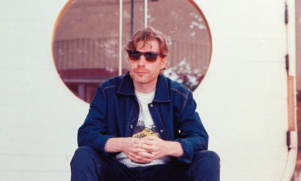 A man wearing sunglasses sits on wooden steps in front of a circular doorway, dressed in a casual outfit.