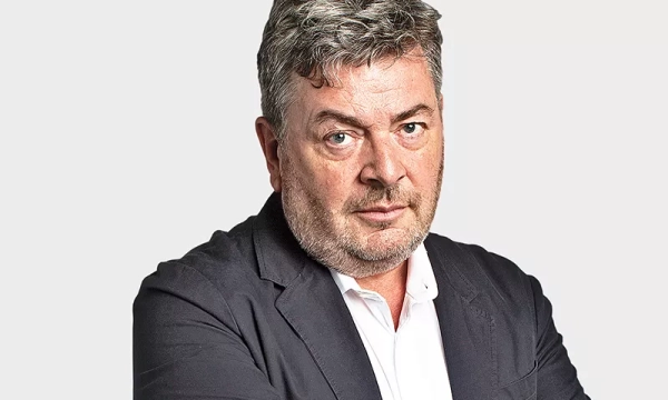 A middle-aged man with gray hair and stubble, wearing a dark blazer over a white shirt, looking directly at the camera with a serious expression.