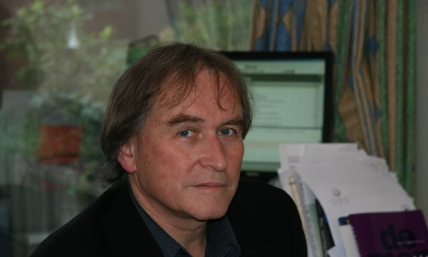 A middle-aged man with dark hair, wearing a black jacket, sits at a desk cluttered with papers, in front of a computer. Natural light filters through a window behind him.