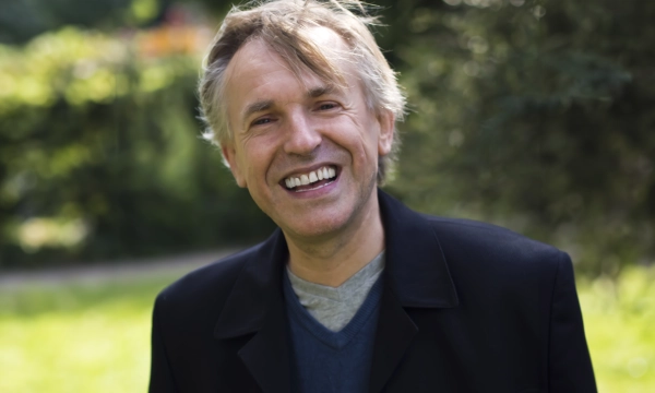 A smiling middle-aged man with gray hair, wearing a black jacket and blue shirt, enjoys a sunny day in a lush green park.