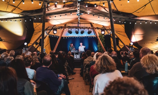 A large audience seated indoors under a tent with warm lighting, watching two speakers at a podium during an event or presentation.