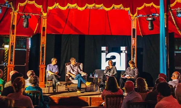 A panel discussion featuring four speakers seated on stage, with an audience in a tent-like venue, decorated in red and blue.