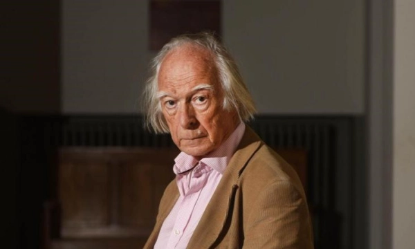 A distinguished elderly man with unkempt gray hair, dressed in a beige jacket and pink shirt, sits thoughtfully in a dimly lit room.