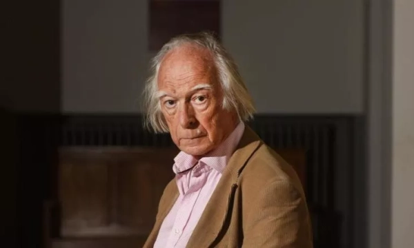 An elderly man with unruly white hair and a serious expression, wearing a beige jacket over a pink shirt, sitting in a dimly lit interior.